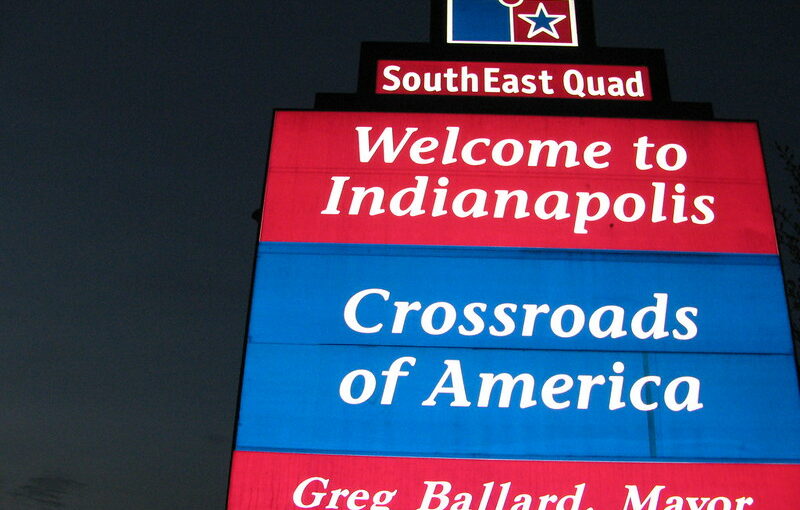 A brightly lit sign saying 'Welcome to Indianapolis Crossroads of America, Greg Ballard, Mayor' against the night sky