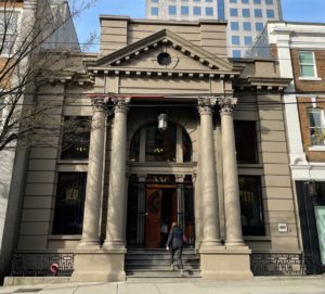 A light brown historical building with columns and steps. The building is small but grand. A modern skyscraper looms in the background.