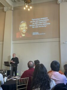 A man presenting in a fancy old building. On a slide above, a quote and photo of a woman: "Technology designers are the new policymakers; we didn't elect them but their decisions determine the rules we live by." Latanya Sweeney Harvard University Director of the Public Interest Tech Lab